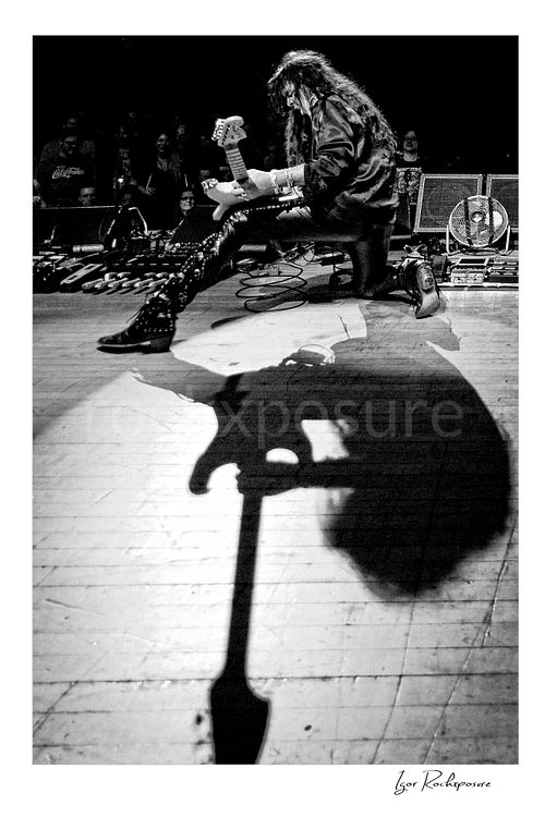 Vertical black and white image of Yngwie Malmsteen in a remote camera shot from the drum kit, kneeling on stage with his guitar while a dramatic shadow stretches across the floor