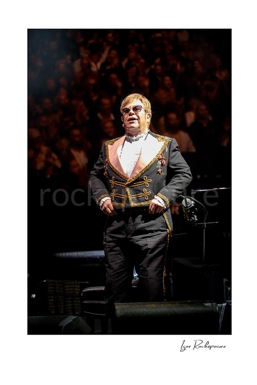 Vertical color image of Elton John standing on stage before a blurred concert crowd in an ornate performance suit