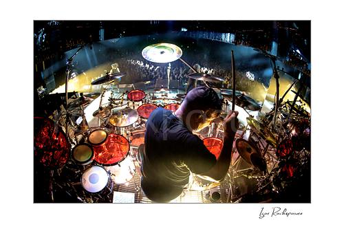 Horizontal color image of Korn drummer Ray Luzier seen from behind at the drums with one drumstick raised above the stage and crowd
