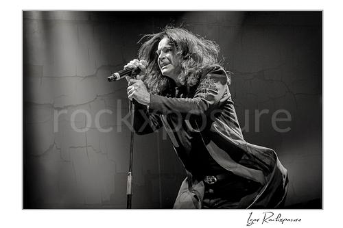 Black and white image of Ozzy Osbourne of Black Sabbath leaning forward singing into a microphone under dramatic stage lighting