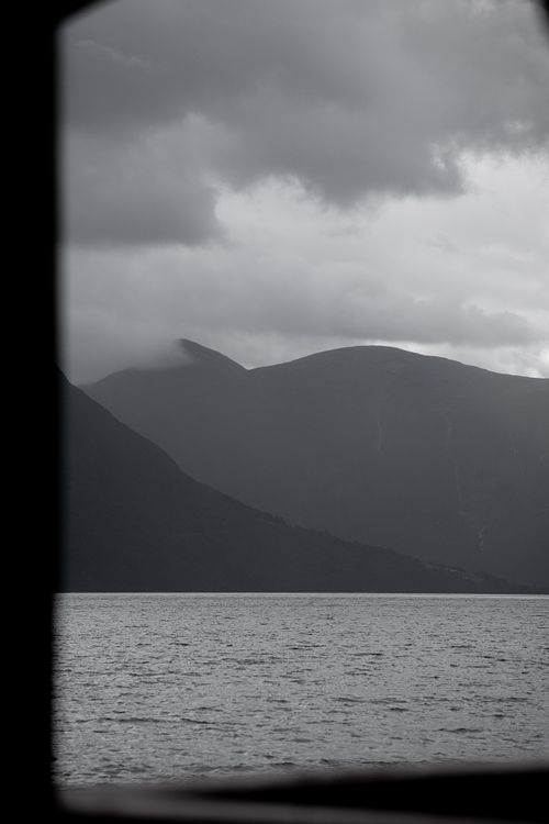 Fjord et montagnes norvégiennes en noir et blanc, sous un ciel chargé, observés à travers un cadre sombre.