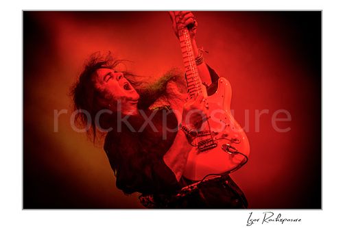 Horizontal color image of Yngwie Malmsteen performing live with a white guitar in a dramatic red-lit stage scene, hair flying and dense stage haze surrounding him