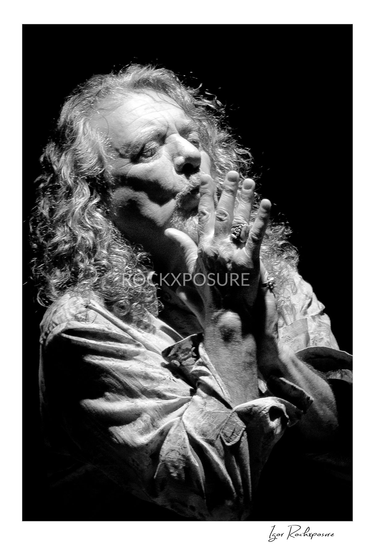 Vertical black and white concert photography of Robert Plant with hands raised near his face in an expressive dramatic portrait under stage lighting