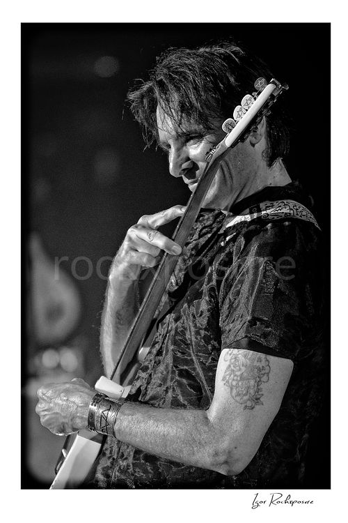 Vertical black and white image of Steve Vai playing guitar in close-up with a playful expression and finger pointed toward the fretboard