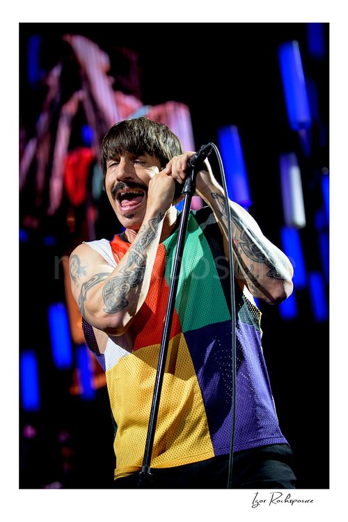 Vertical color image of Anthony Kiedis of Red Hot Chili Peppers singing at the microphone in a tight live performance close-up, with tattooed arms and glowing blue LED tubes behind him