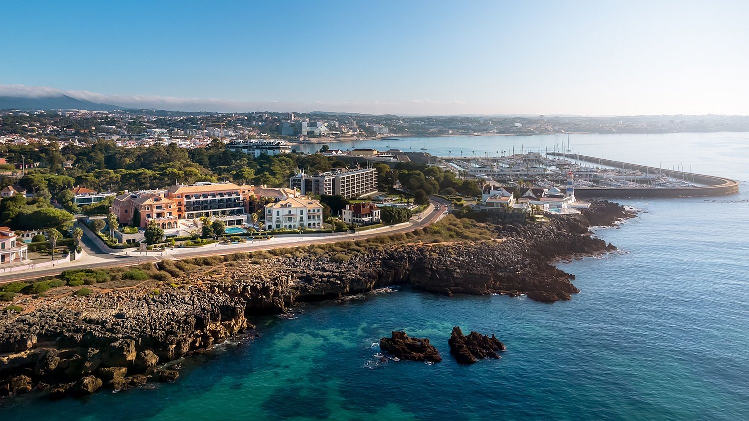 Vista a&eacute;rea do Grande Real Villa It&aacute;lia Hotel & Spa em Cascais, membro dos Leading Hotels of the World, com a marina e o farol de Santa Marta ao fundo, fotografada por Pedro Br&aacute;s Photography &mdash; fotografia a&eacute;rea profissional de hotelaria e resorts de luxo e