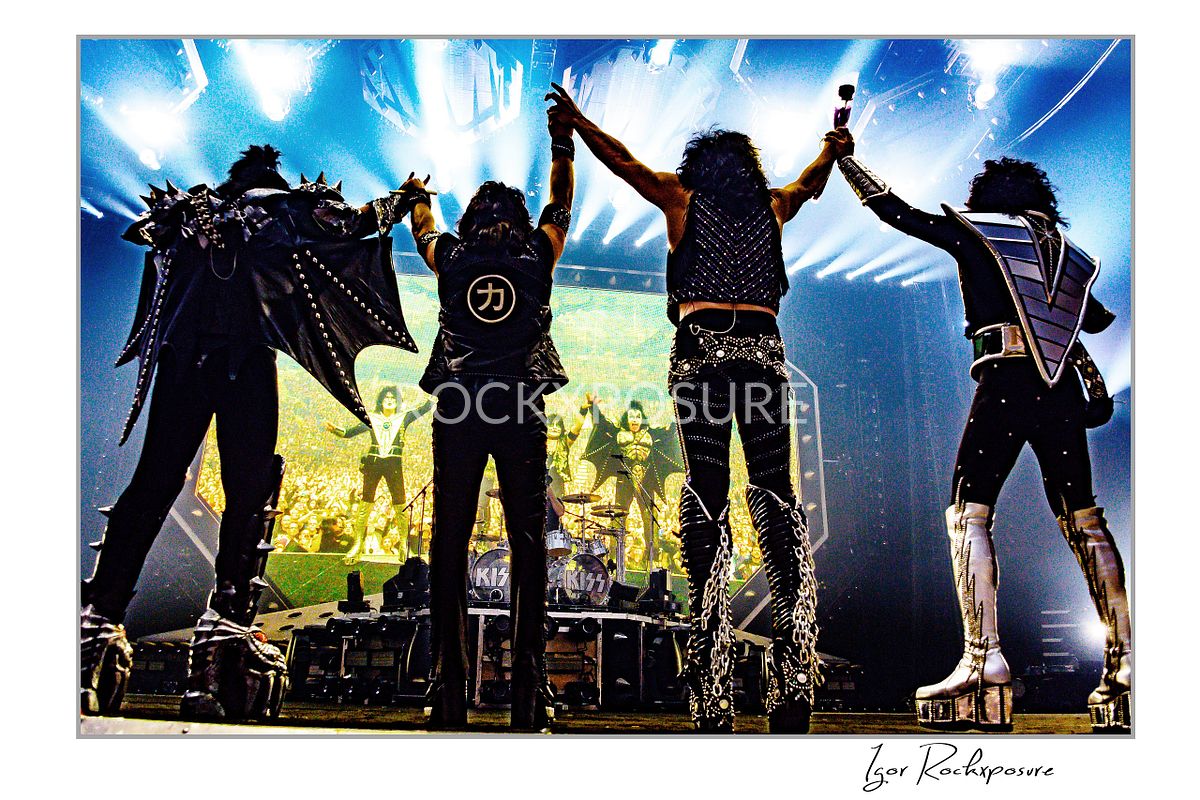 Horizontal color photograph of the KISS band standing onstage with their backs to the camera holding hands in the air while facing a massive stage screen that displays their own image, creating a dramatic mirrored moment in front of the crowd