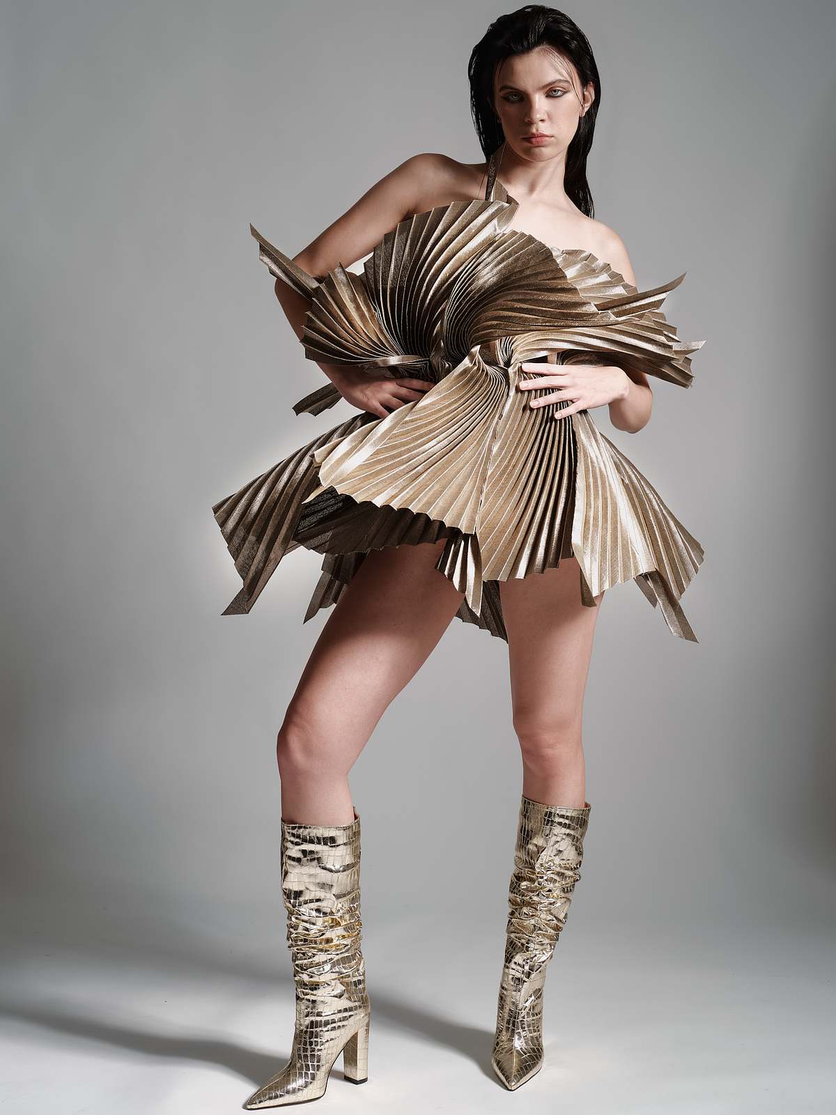 A woman stands with her right leg placed ahead of her left leg, her hands on her hips and is wearing a gold colored pleated  dress with a sculptural silhouette and golden knee length boots with heels captured full length in the photo