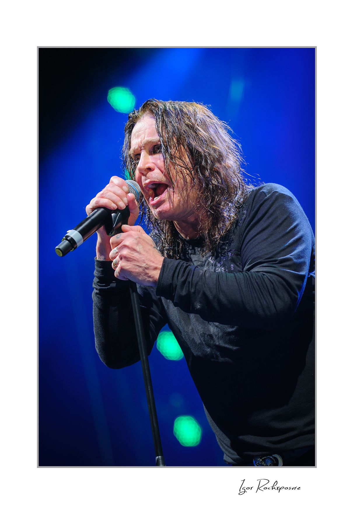 Ozzy Osbourne of Black Sabbath singing into a microphone during a live performance under deep blue concert lighting