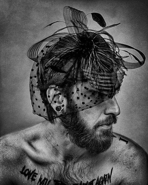 Portrait en noir et blanc d’un homme barbu et tatoué portant une voilette délicate, exprimant la tendresse masculine assumée et la liberté d’identité — photographie fine art par Pascal Yennis, Namur, Belgique.