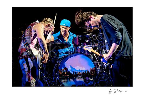 Horizontal color image of Flea, Chad Smith, and Josh Klinghoffer of Red Hot Chili Peppers performing live around the drum kit, with Smith striking the drums while Flea and Klinghoffer lean in on either side