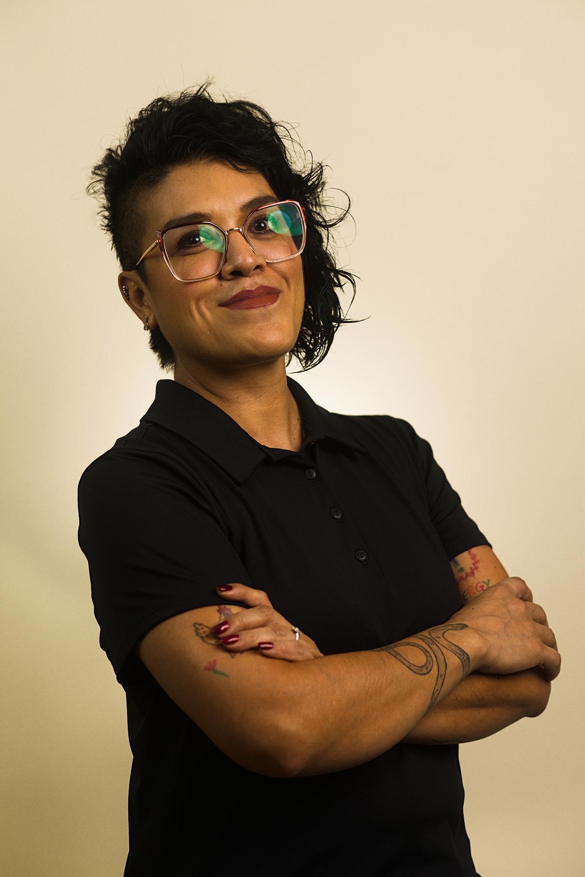 Retrato de una mujer profesional, con pose de brazos cruzados y sonriente, porta algunos tatuajes que son visibles, los cuales añaden un aire de confianza, familiaridad y transparencia.