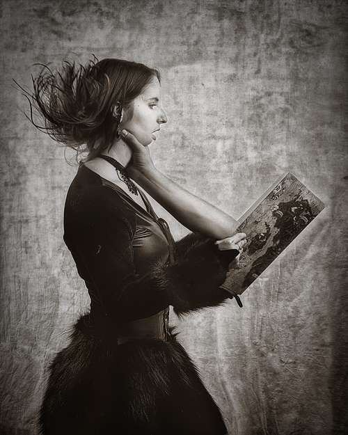 Portrait artistique en noir et blanc d&rsquo;une femme en costume tenant un livre dont une main semble &eacute;merger, la saisissant &agrave; la gorge, &eacute;voquant une absorption surr&eacute;aliste par la fiction &mdash; photographie fine art par Pascal Yennis, Namur, Belgique.