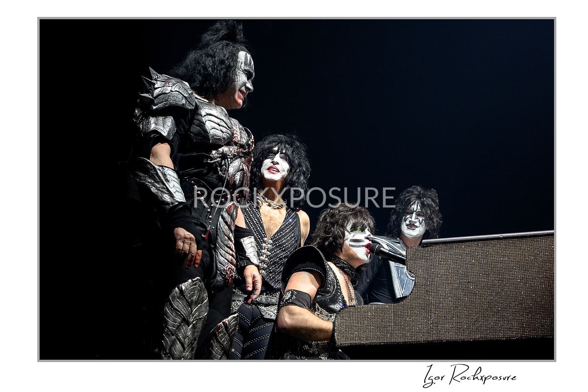 Horizontal color photograph of the full KISS band in full makeup and costumes gathered closely around Eric Singer as he plays piano and sings in a theatrical staged moment