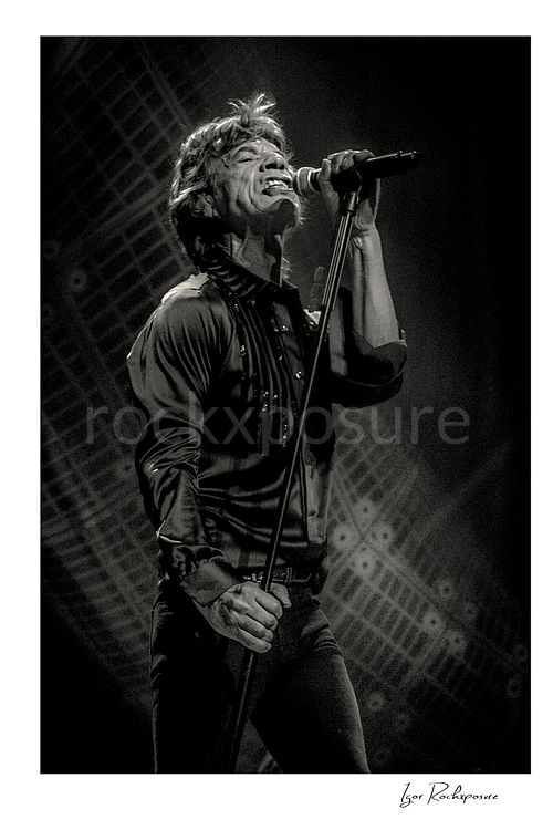 Vertical black and white image of Mick Jagger of The Rolling Stones singing into a microphone in a dramatic stage portrait