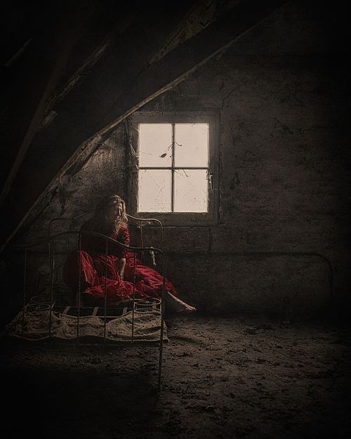 Portrait artistique d'une femme en robe rouge assise sur un lit ancien dans un grenier sombre, évoquant l’abandon, la solitude et la reconstruction intérieure — photographie fine art par Pascal Yennis, Namur, Belgique.