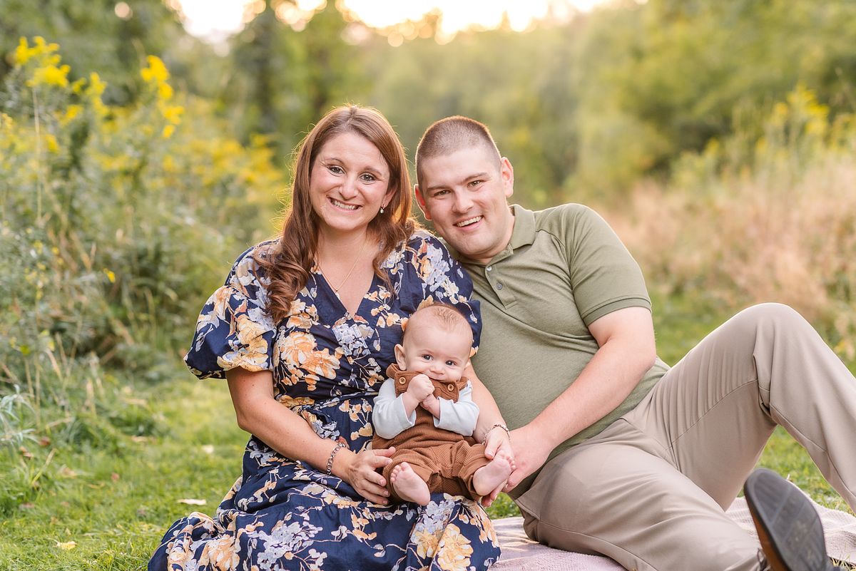 Husband and wife sitting in a green field, holding their new baby and smiling