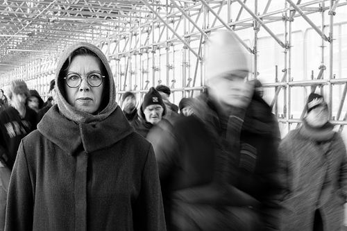 Kvinna i kappa står stilla medan människor passerar suddigt i byggställningstunnel