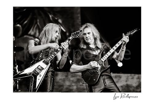 Horizontal black and white image of Richie Faulkner and Glenn Tipton of Judas Priest performing live on stage, with Faulkner at left playing flying V guitar and Tipton at right playing electric guitar