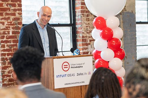 Intuit executive speaking at a press conference for the IDEAS program, in partnership with Urban League of Philadelphia, addressing attendees in front of branded signage and balloons, captured by professional event photographer.