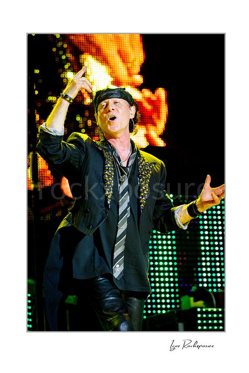 Vertical color image of Klaus Meine of Scorpions performing live in cap and embellished jacket with glowing orange and green stage screens