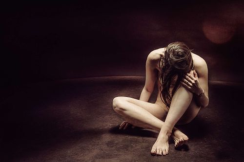 Femme nue assise au sol dans un d&eacute;cor sombre, pench&eacute;e vers l&rsquo;avant, symbolisant la culpabilit&eacute; et les cons&eacute;quences du karma
