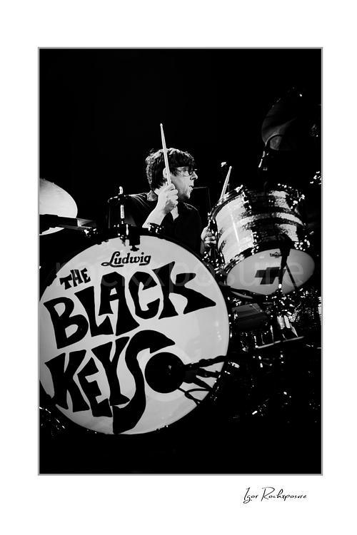 Vertical black and white image of Patrick Carney of The Black Keys playing drums beside a bass drum marked The Black Keys