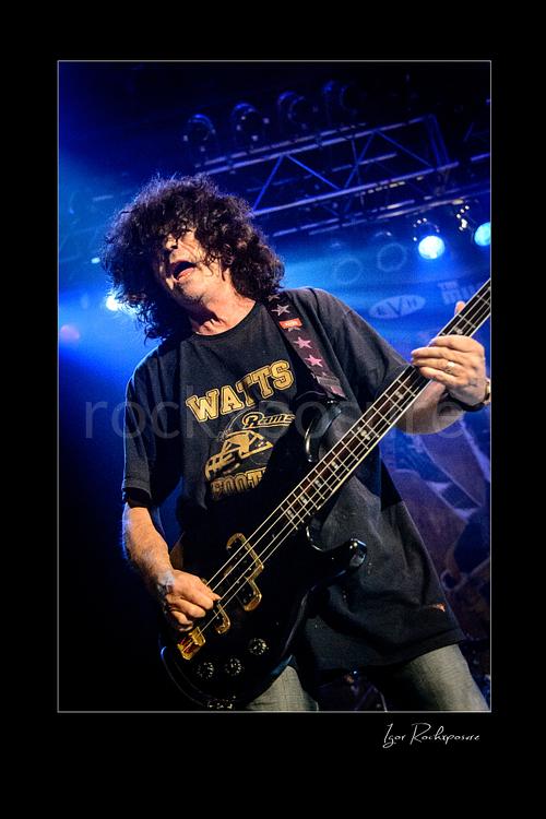 Vertical color image of Jimmy Bain playing bass onstage under blue concert lighting in an energetic live performance shot
