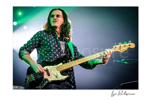 Horizontal color image of Geddy Lee performing with his bass guitar during a Rush concert under green stage light