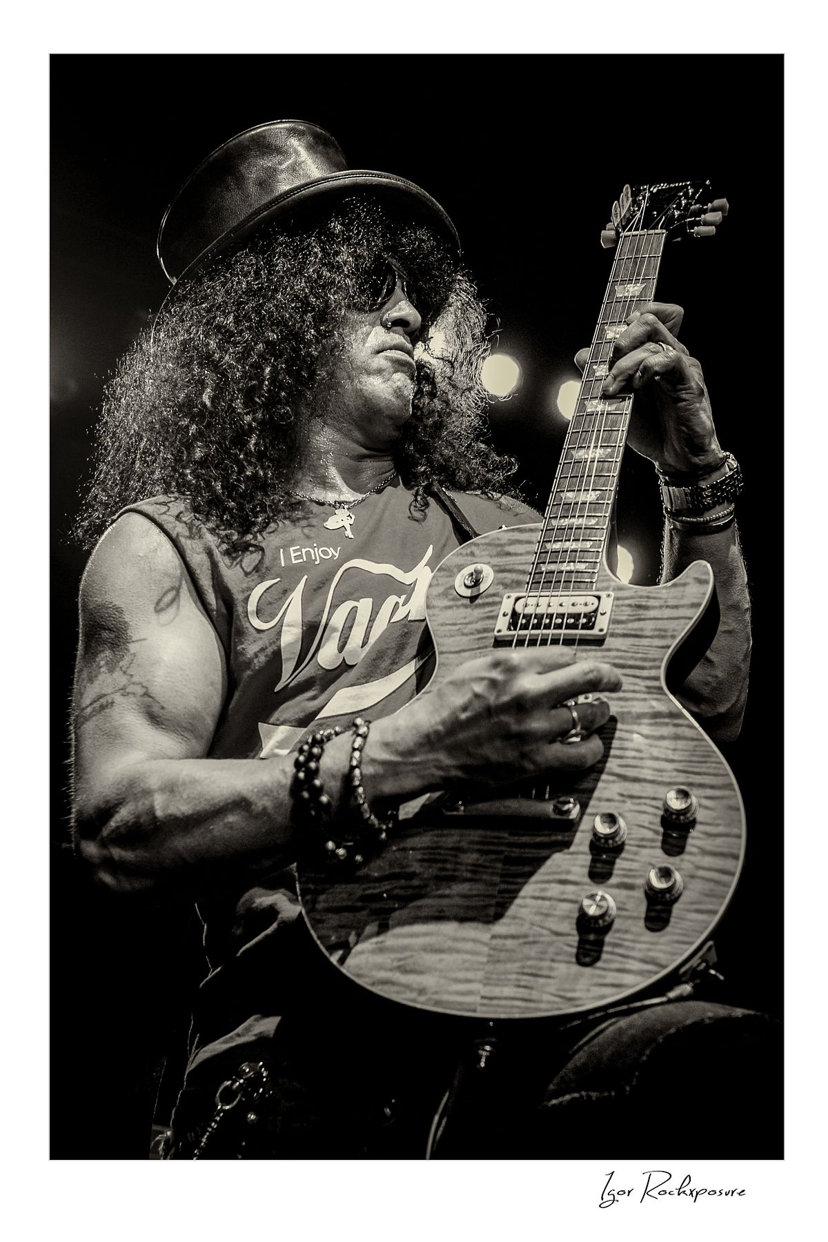 Vertical concert photography of Slash playing electric guitar in black and white photography wearing his signature top hat under dramatic stage lighting