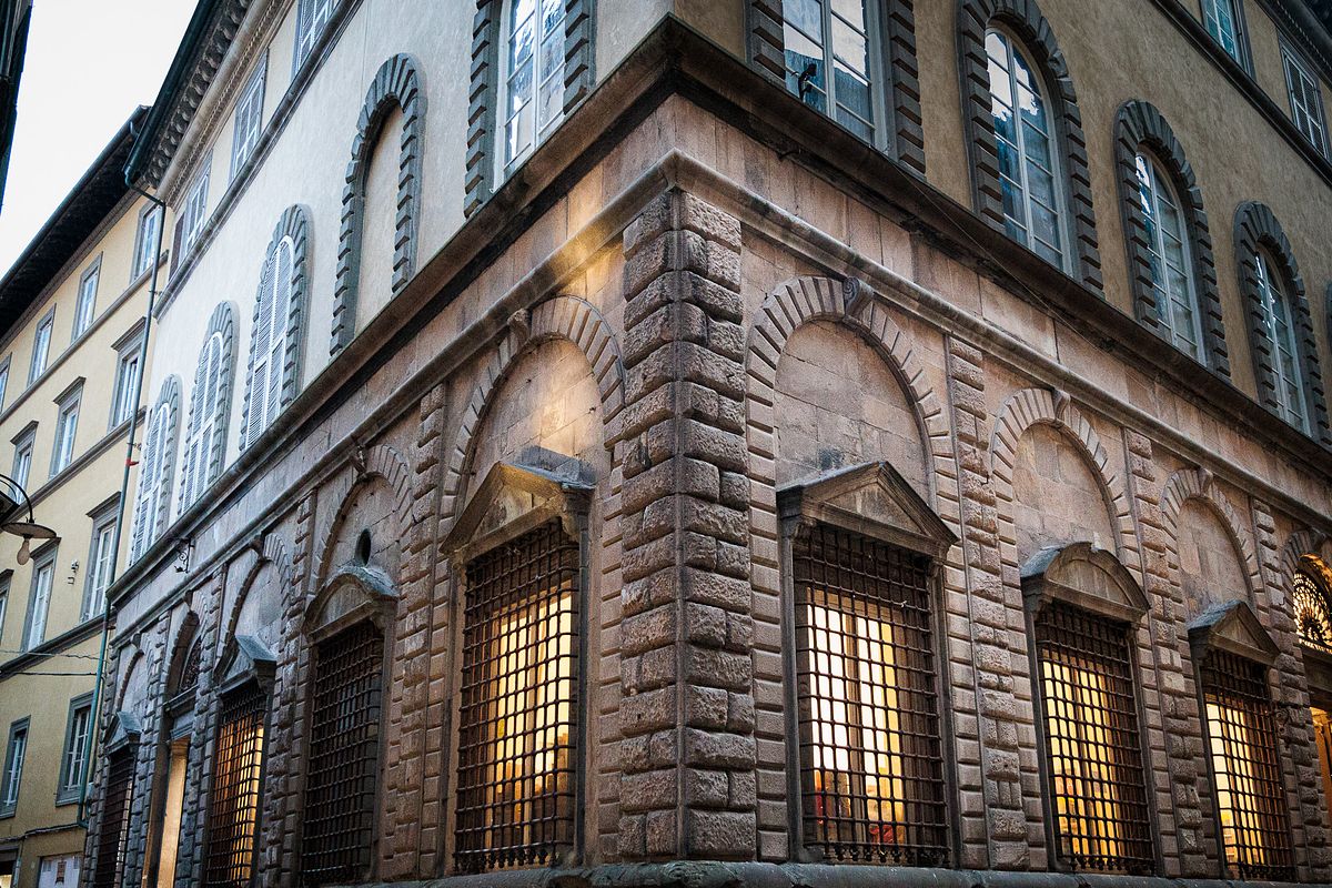 Evening street scene in Lucca, Italy, showing the corner of a historic stone building with two large arched windows covered by metal grilles glowing warmly from interior light, smaller windows above and several people standing in the foreground, creating a moody blue-hour atmosphere.