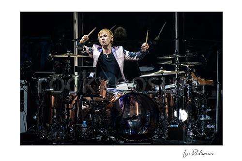 Horizontal color image of Dominic Howard of Muse at the drum kit with both drumsticks raised in a dramatic live performance