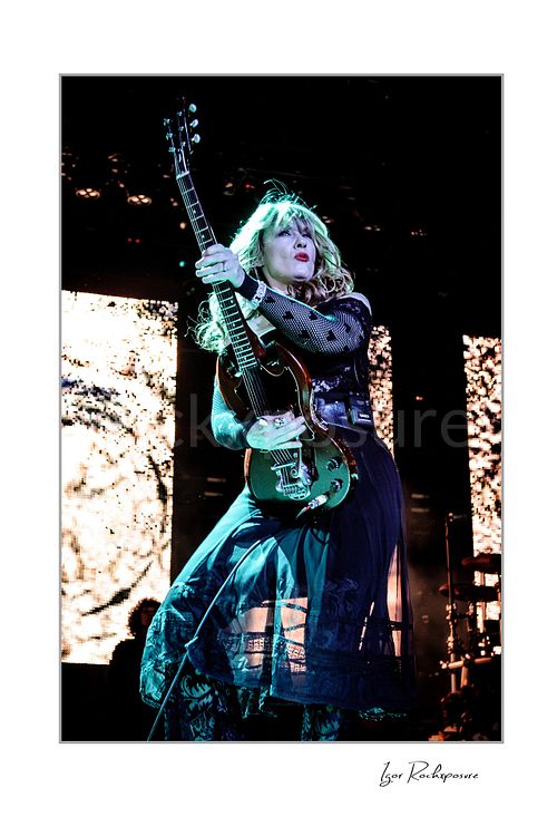 Vertical color image of Nancy Wilson performing with electric guitar during a Heart concert, framed by bright backlight and flowing stagewear