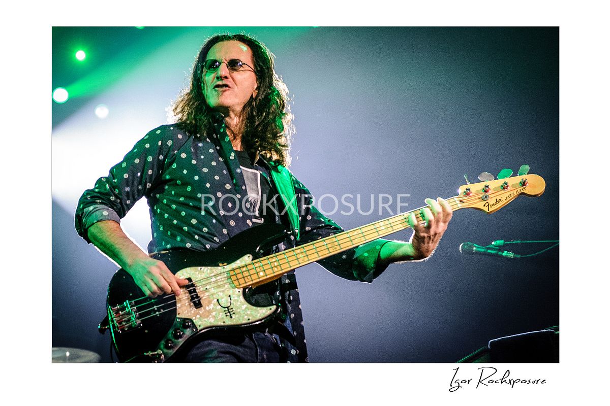 Horizontal concert photography of Geddy Lee playing bass guitar under green stage lighting