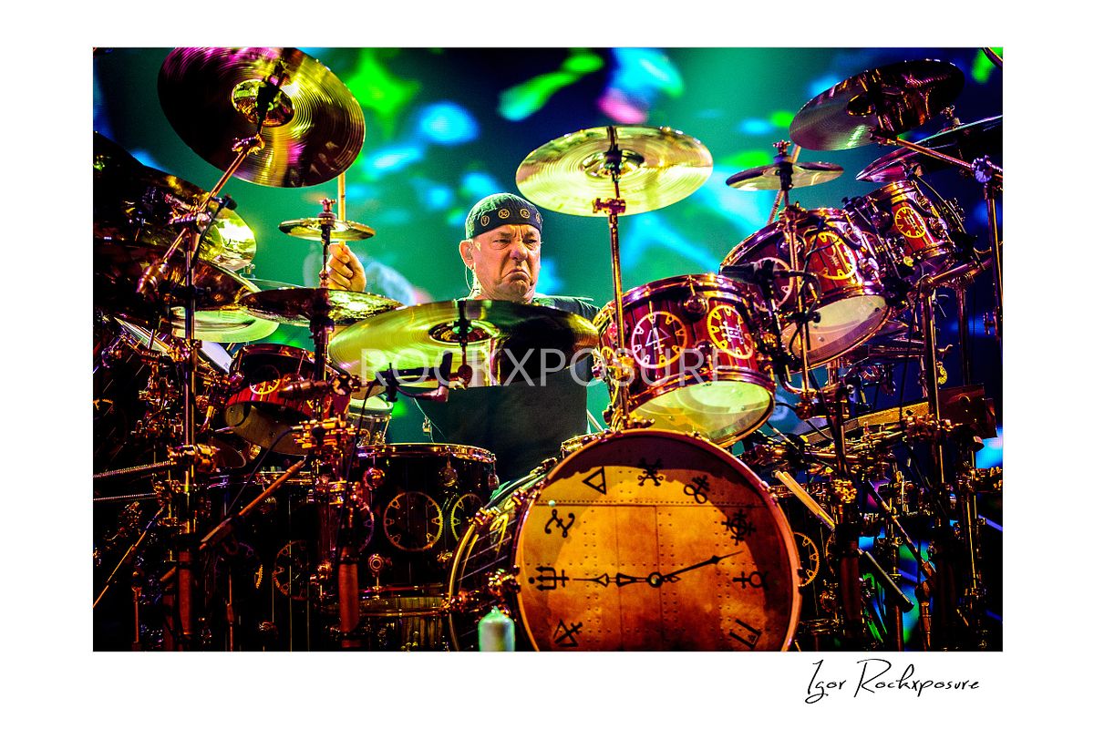 Horizontal concert photography of Neil Peart performing behind a large drum kit under colorful stage lighting