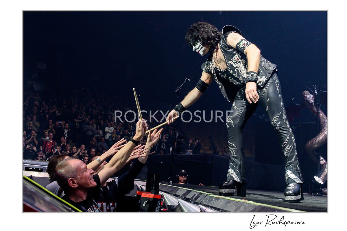 Horizontal color photograph of Eric Singer in full KISS makeup and costume leaning forward from the stage to hand his drumsticks to an excited fan in the crowd during a high‑energy concert moment