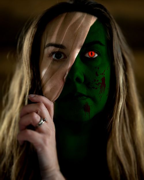 A woman peels the top layer of her face away to reveal a red-eyed green demon beneath.