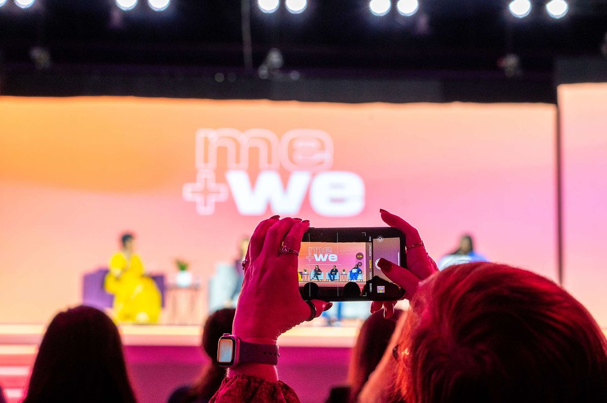 Audience member records “me + we” panel at a corporate event using her smartphone, capturing content for social media.