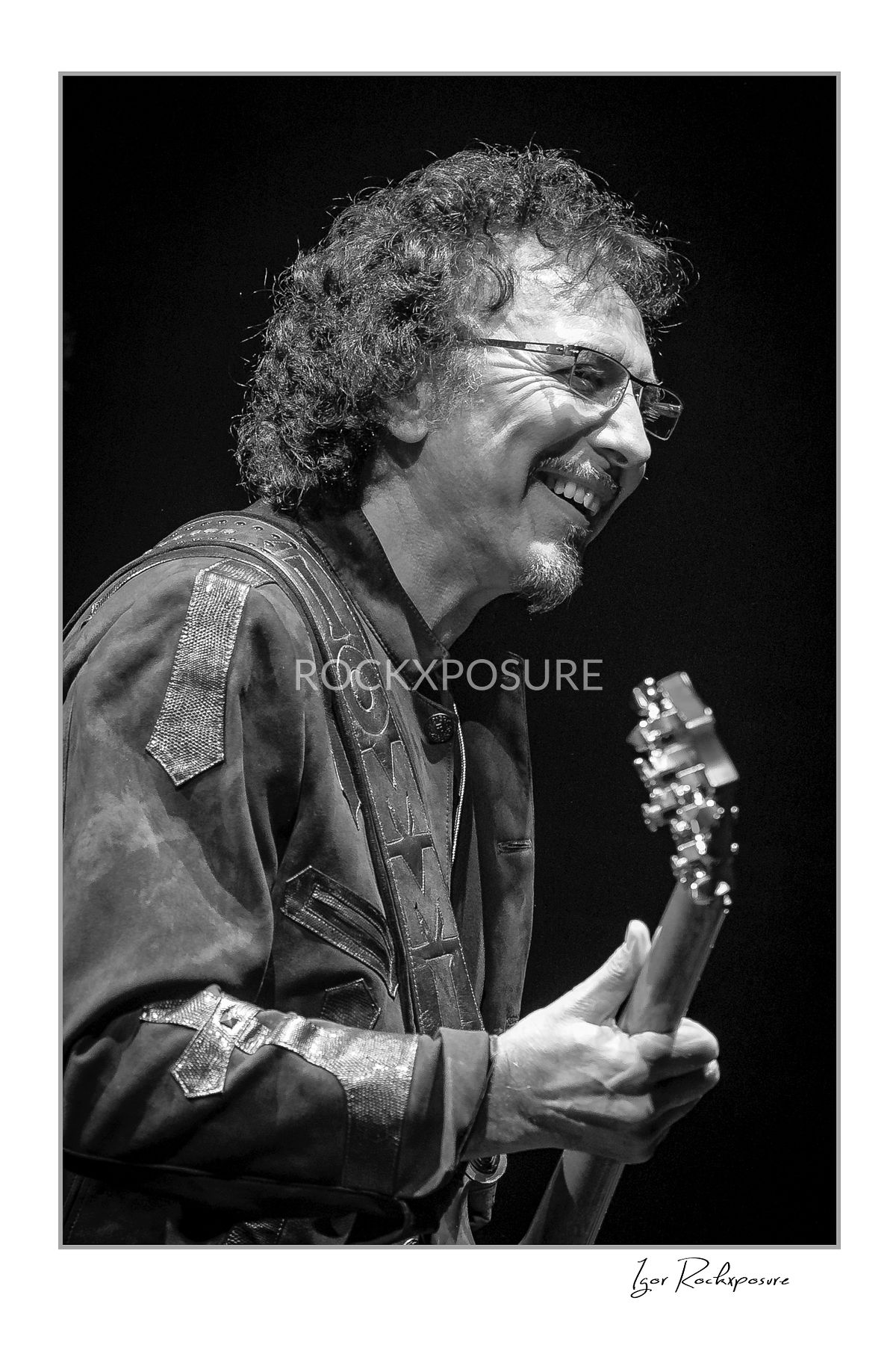 Black and white portrait of Tony Iommi of Black Sabbath smiling while holding his electric guitar during a live performance