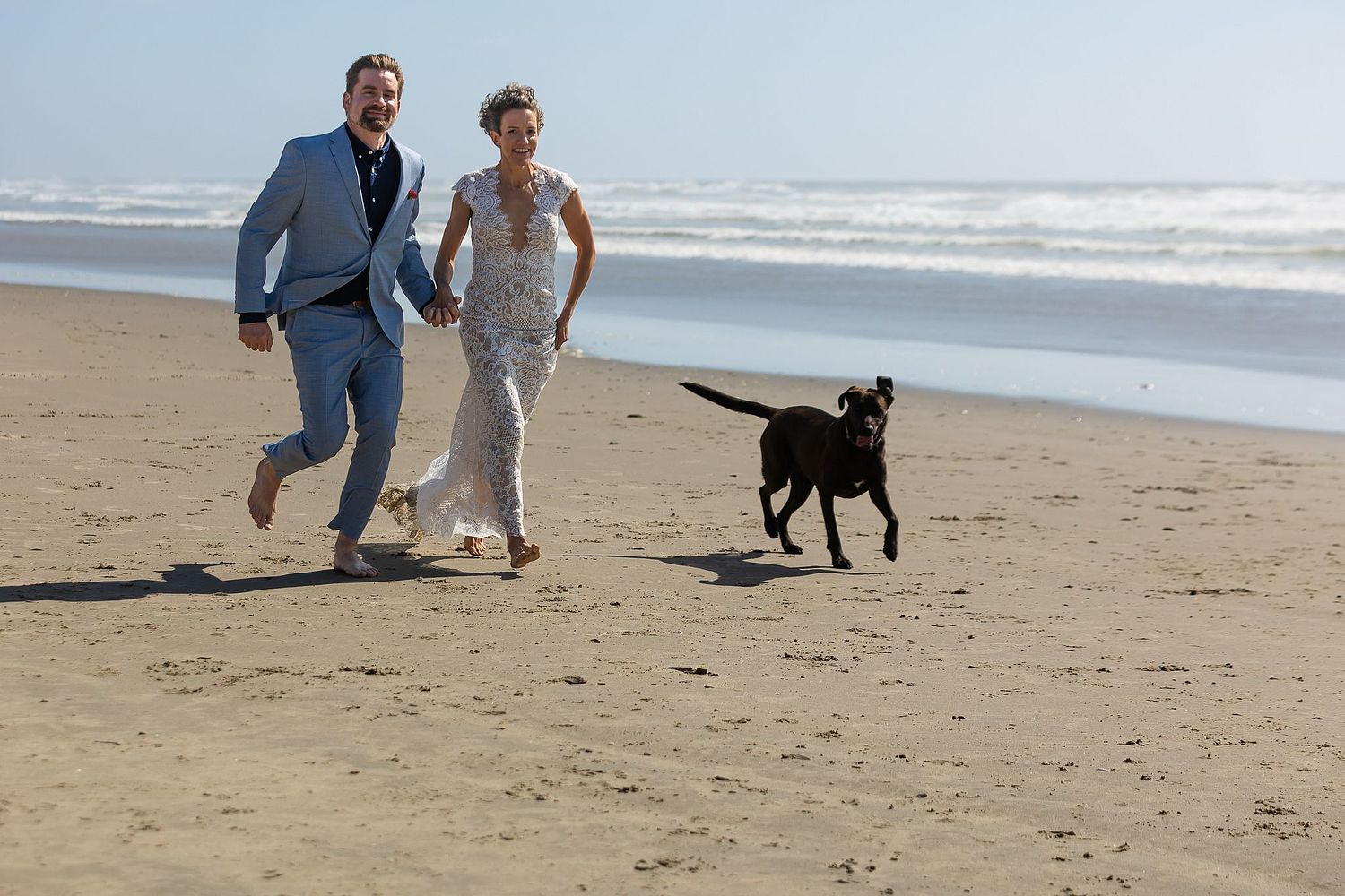 A newly married couple running along the beach with their dog