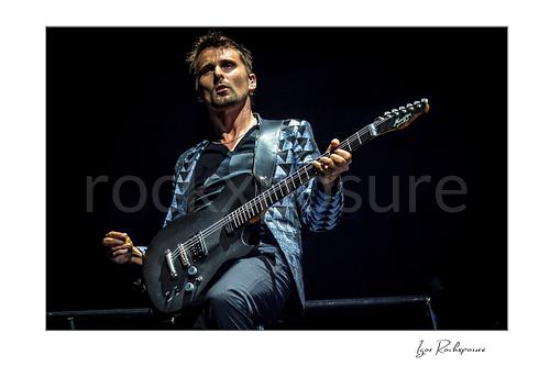 Horizontal color image of Matt Bellamy of Muse playing guitar in a patterned stage jacket against a dark concert background